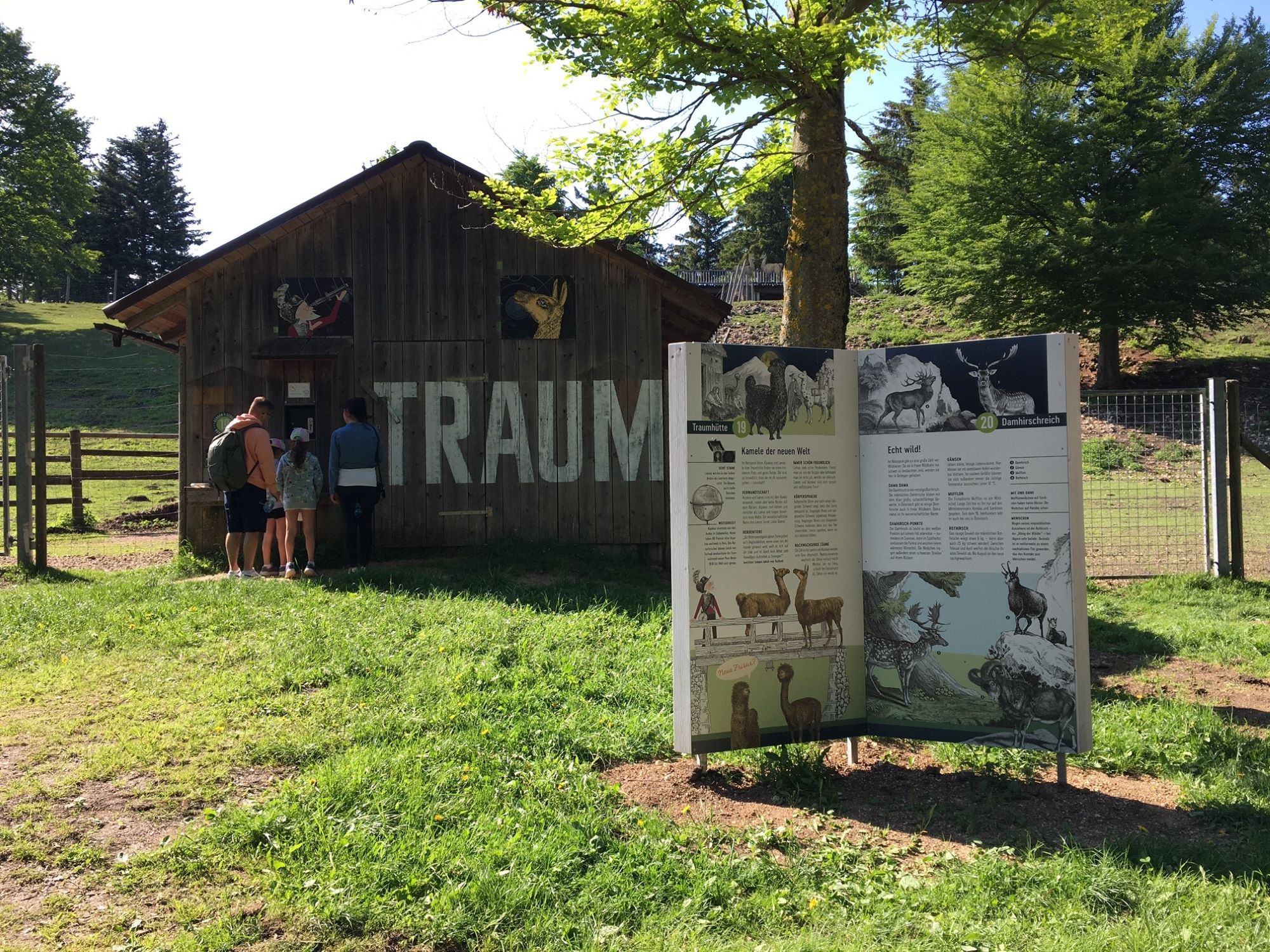 A family stands in front of a wooden hut with the inscription 'TRAUM'. Next to it is an information board about animals in a green park.