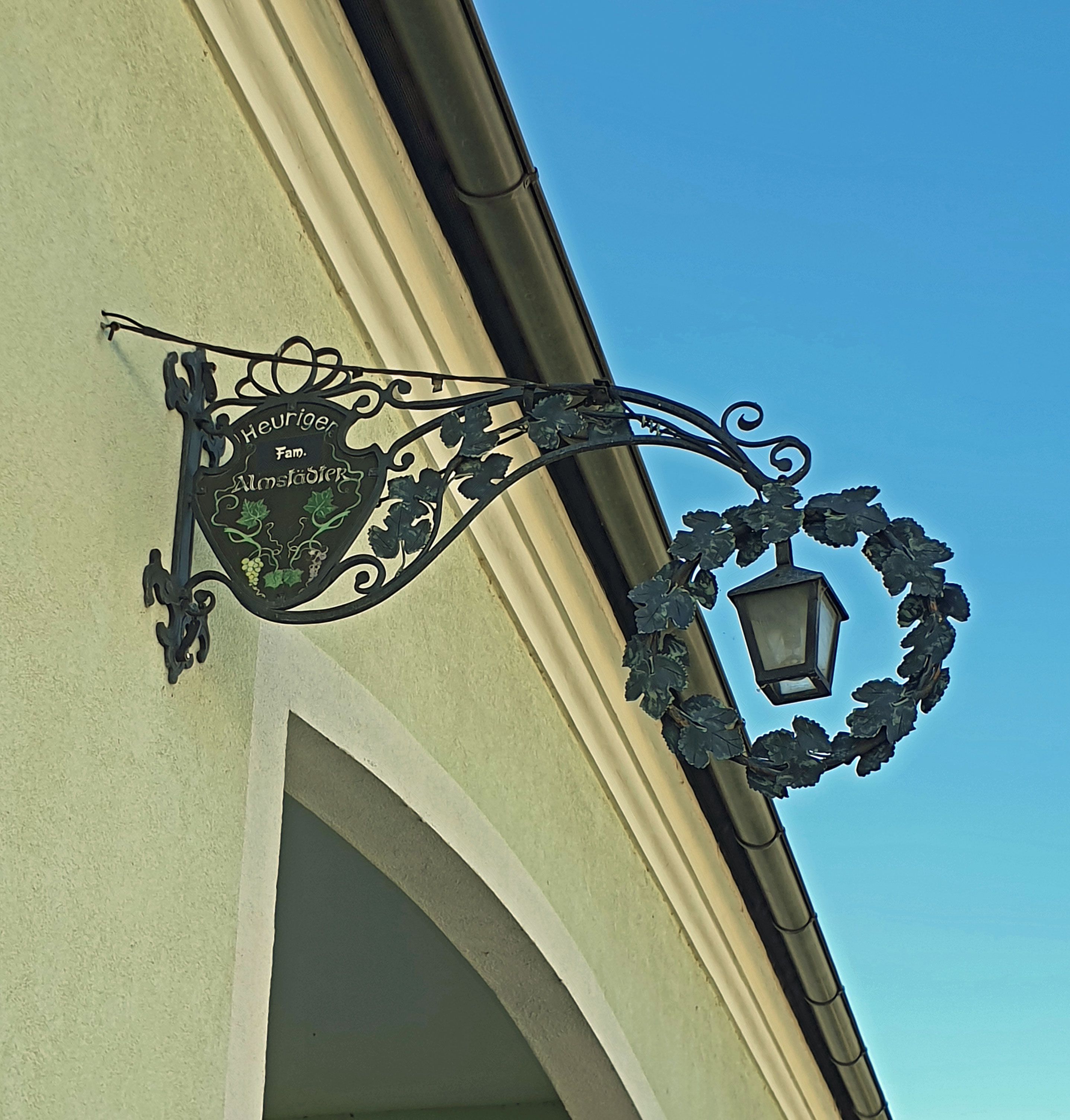 Metal sign with lantern on a house wall, labeled 'Heuriger Fam. Almstädter'.