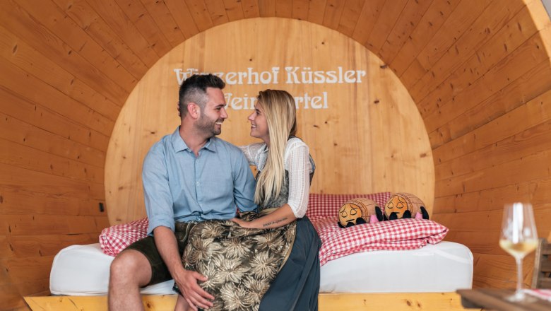 A couple is sitting in a large wooden wine barrel that has been converted into a suite.