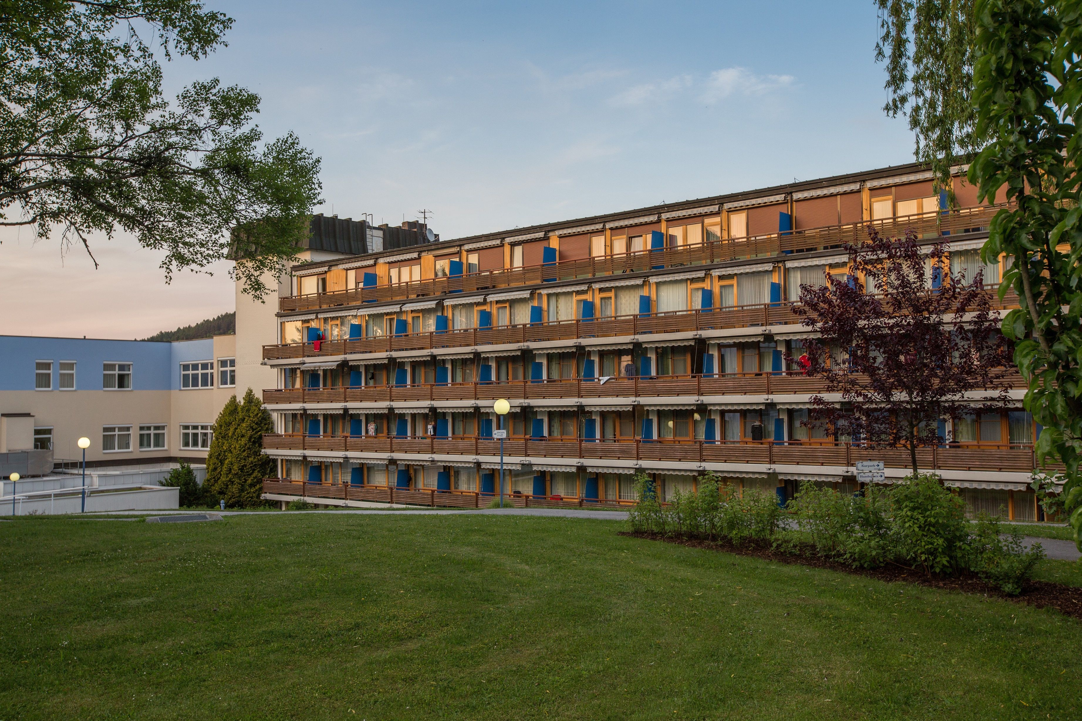 A multi-storey building with balconies and blue awnings, surrounded by trees and lawns.