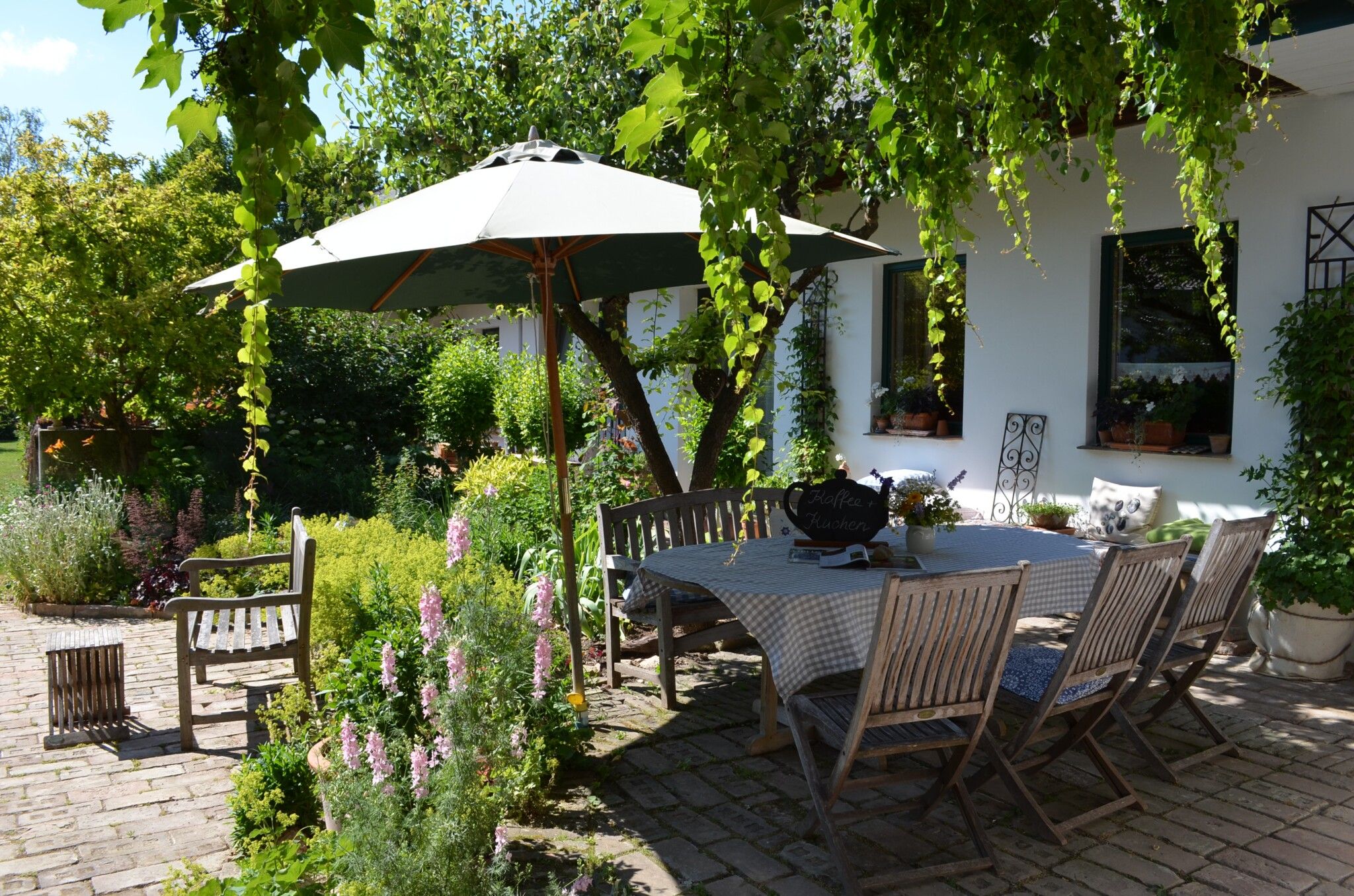 An idyllic garden with a wooden table and chairs under a parasol, surrounded by green plants and flowers.