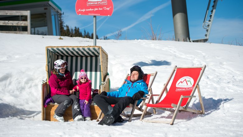 Family relaxing in the snow on the Erlebnisalm, sitting in a beach chair and deckchairs under a sign saying 'Genussplatz'.