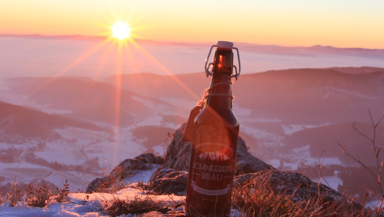 A bottle of Schneeberg Br&auml;u beer stands on a snow-covered mountain at sunset.