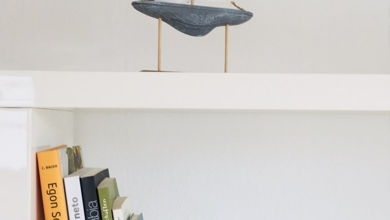 A white shelf with books and a small sailing boat as decoration.