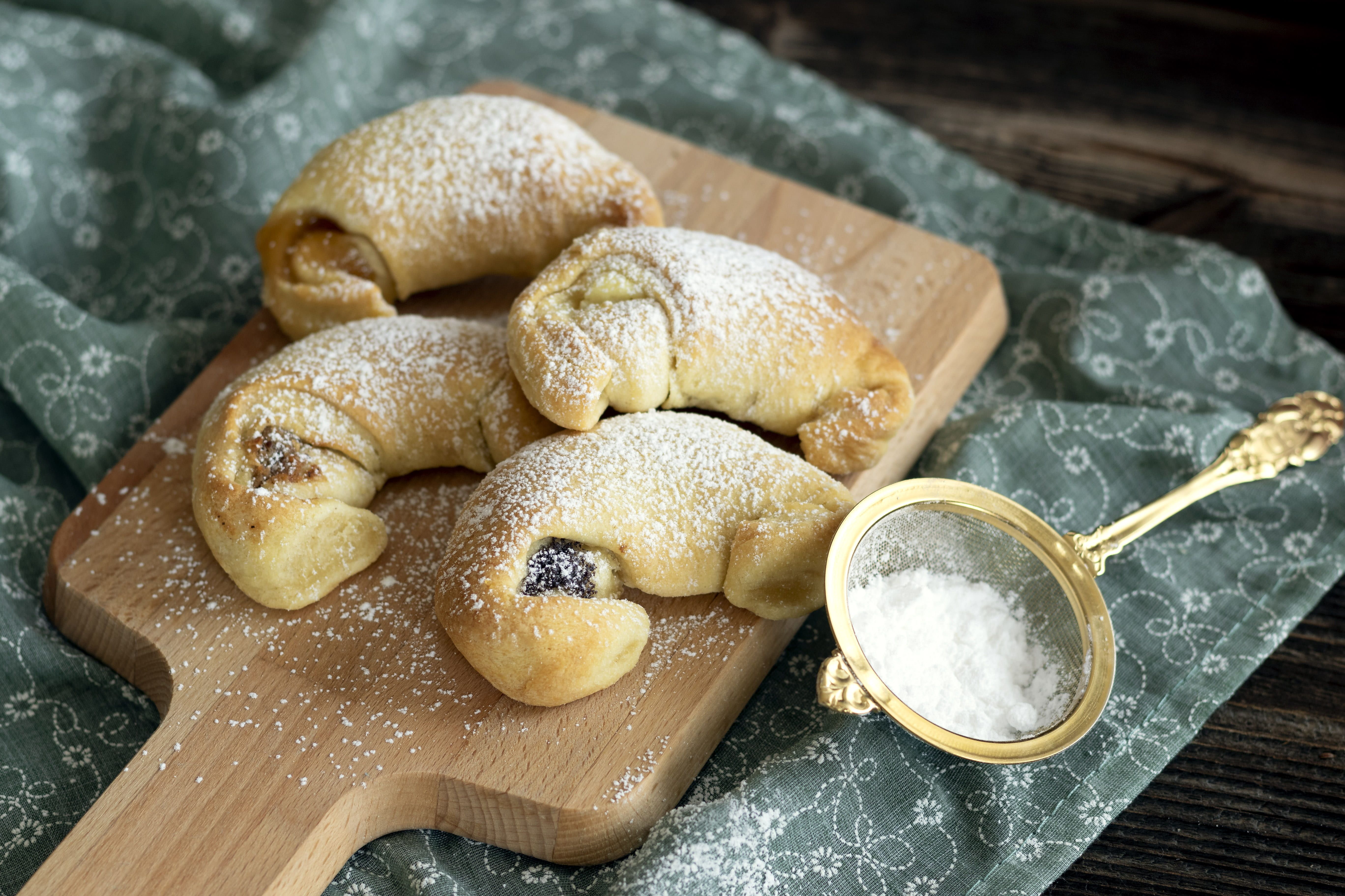 Four cream croissants on a wooden board sprinkled with powdered sugar, next to a sieve with powdered sugar.