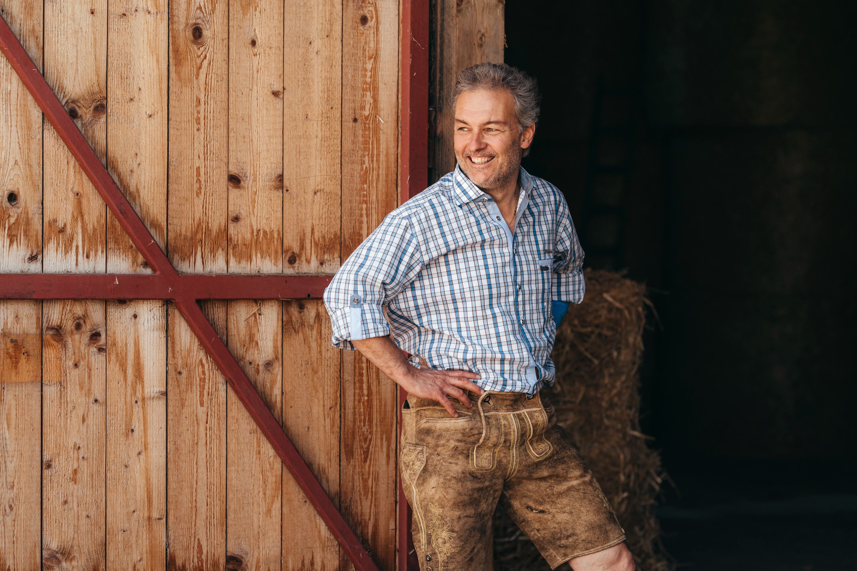 Man in plaid shirt and leather pants leans against wooden wall, smiles.
