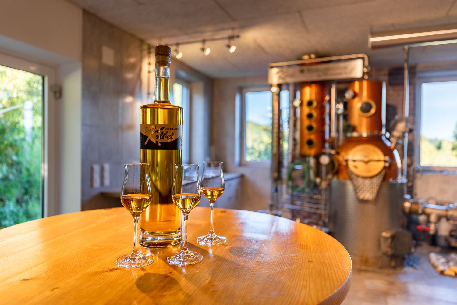 A bottle and three half-filled glasses stand on a round wooden table in front of a still.