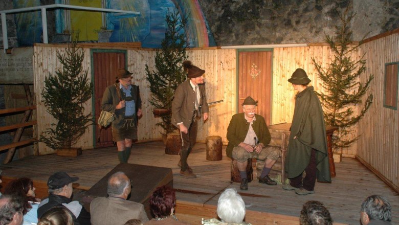 Theater performance with four men in traditional dress on a wooden stage, surrounded by fir trees, in front of an audience.