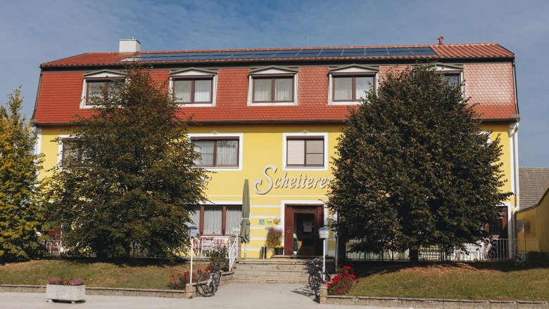 Yellow building with a red roof and the inscription 'Scheiterer', surrounded by trees and bicycles.