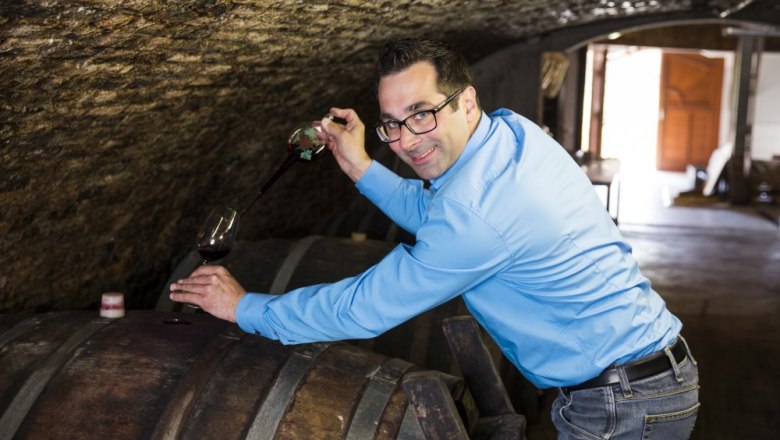 Man in blue shirt draws wine from a barrel in a wine cellar.
