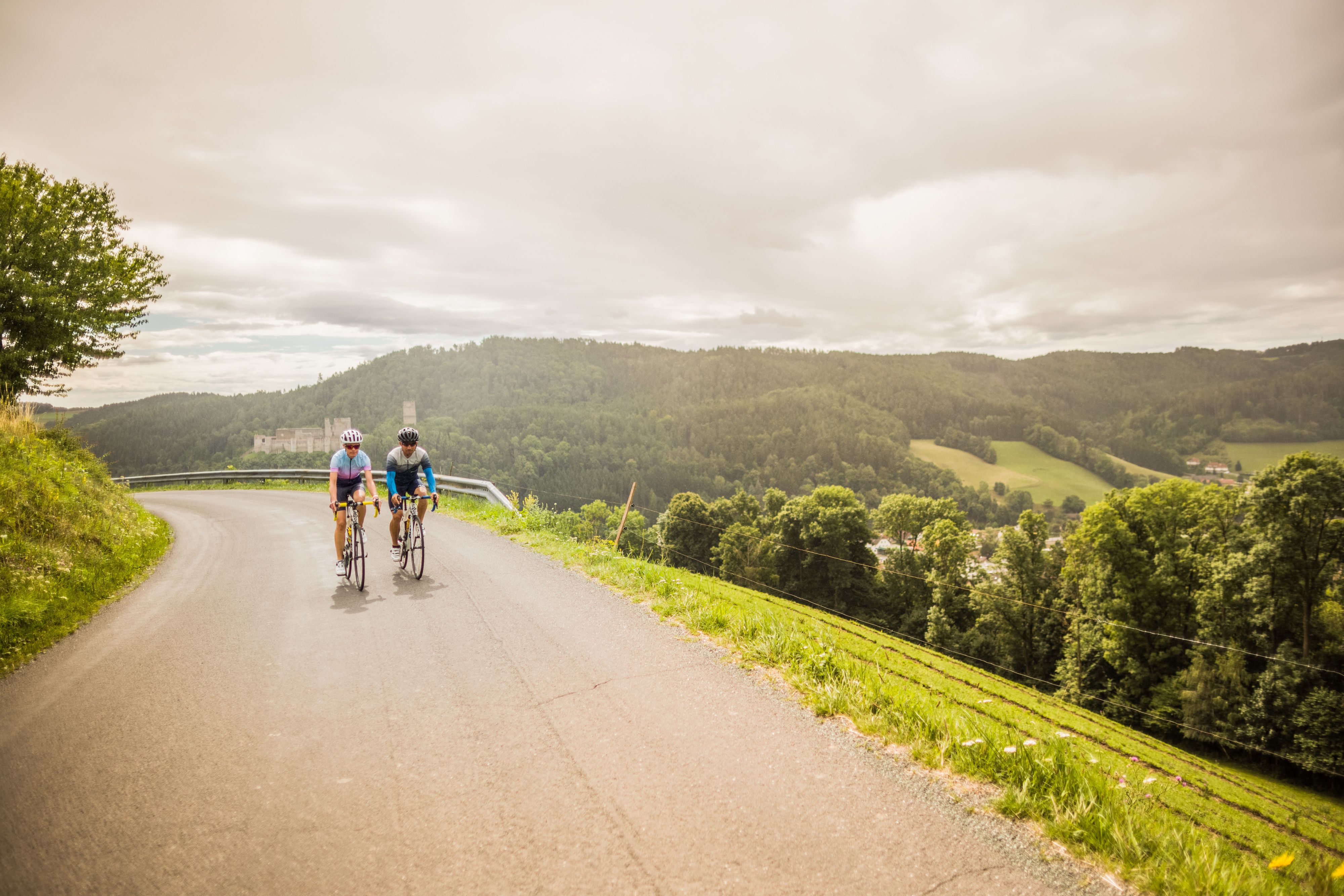 Two people on bicycles on a road in a hilly landscape with a castle in the background.