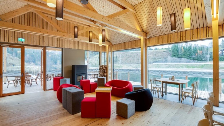 Interior view of a modern room with wooden walls, colorful seating and large windows overlooking a terrace and nature.