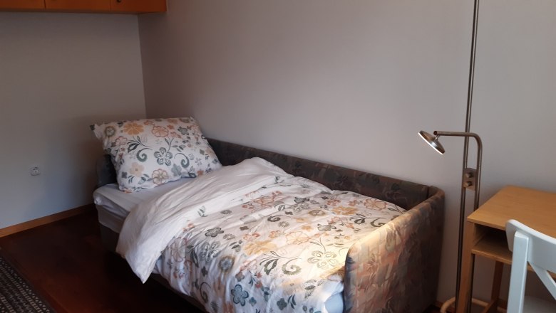 A small room with a single bed, a desk with a lamp and a chair. The bed has colorful floral bedding.