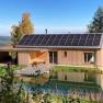 A modern wooden house with solar panels on the roof, surrounded by nature and a small pond in the foreground.