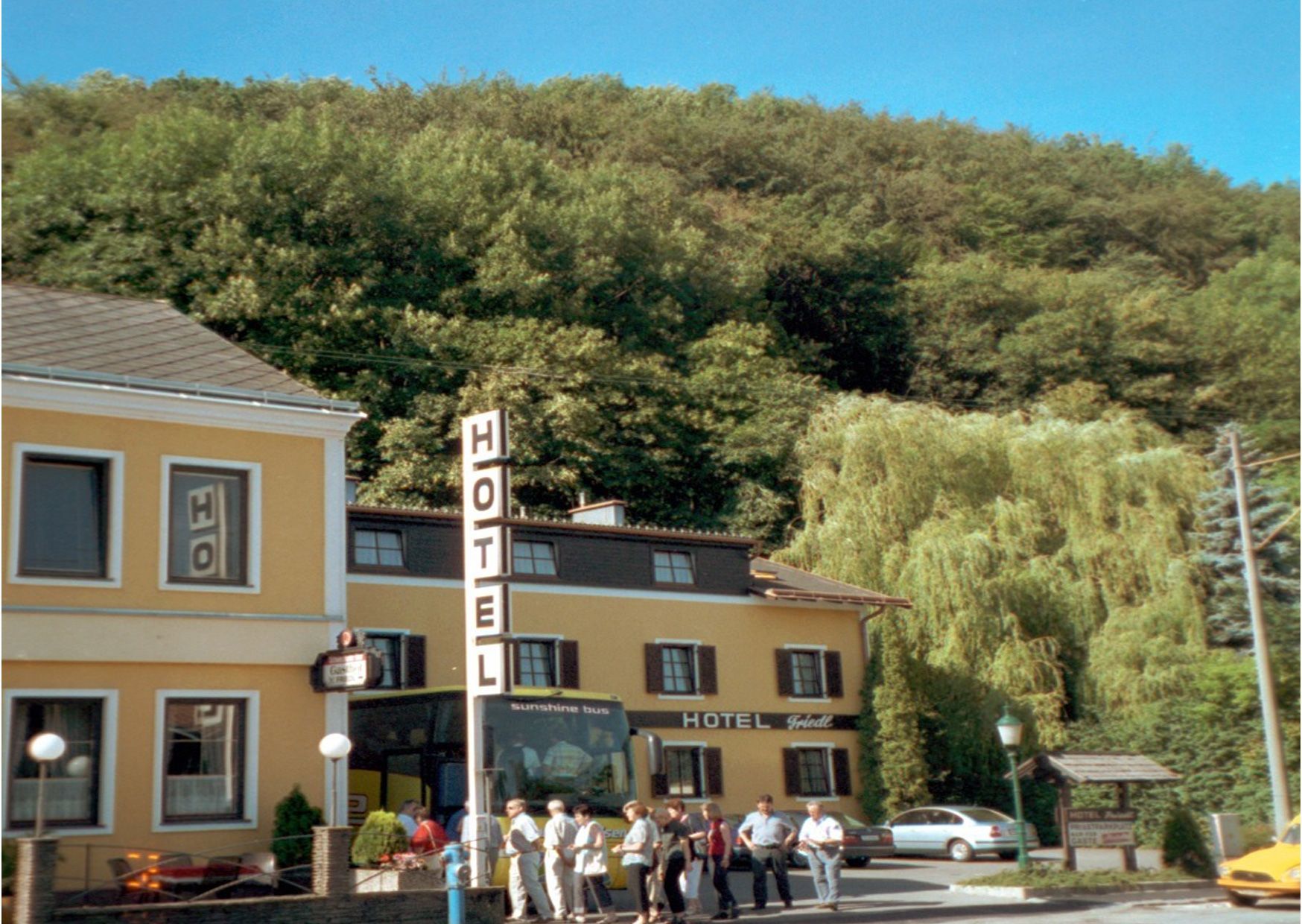 A yellow hotel building with a large hotel sign and people in front of it, surrounded by trees.