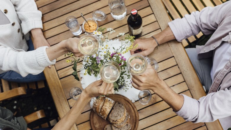 Three people clink glasses of wine at a wooden table with bread and flowers on it.