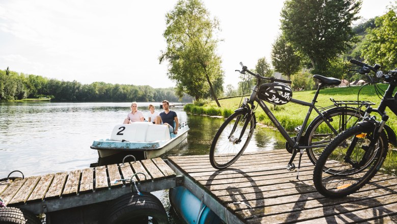 Three people on a pedal boat on a lake, next to them bicycles on a jetty.