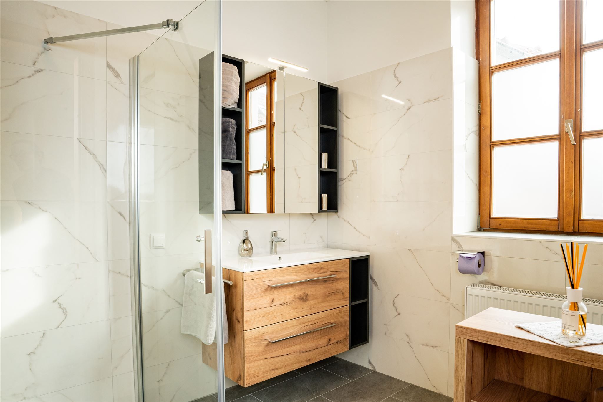 Modern bathroom with wooden furniture, large window front and glass shower.