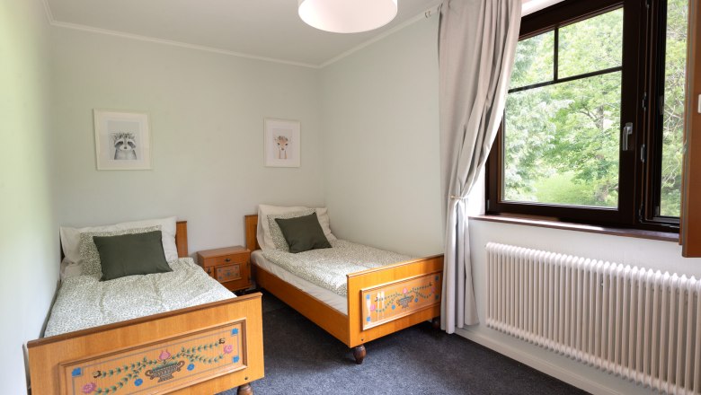 Bedroom with two single beds, wooden furniture and a large window with a view of the greenery.