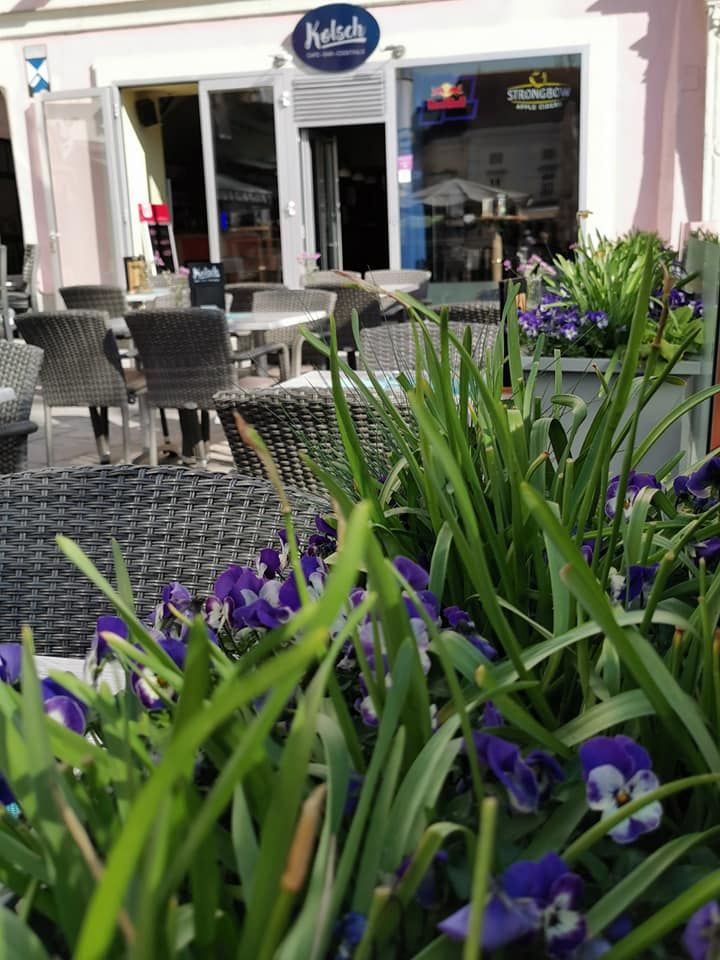 A sidewalk garden with wicker chairs and tables in front of a café, surrounded by blooming flowers and plants.
