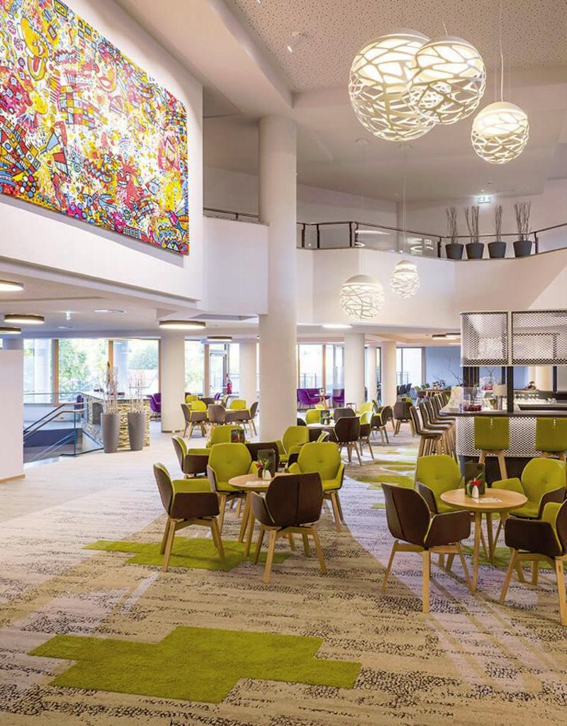 Modern hotel lobby with carpeted floor, round tables, green chairs, large lamps and a mural.