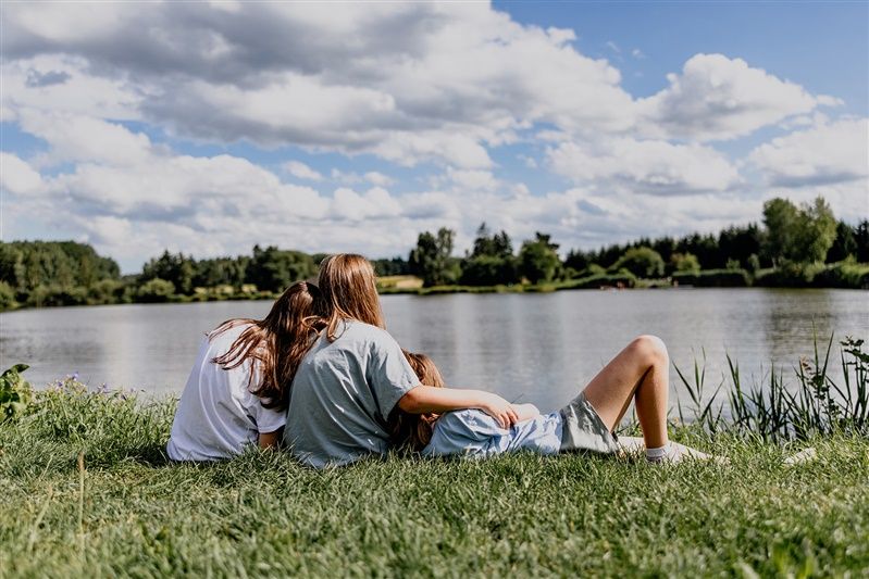 Three people sit on a meadow by the lake and look out over the water.