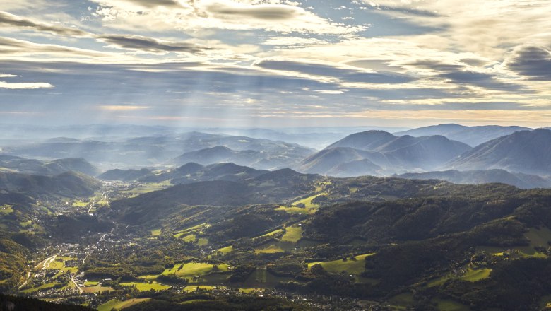 Panoramic view of the Rax Alps with wooded hills and rays of sunshine through clouds.