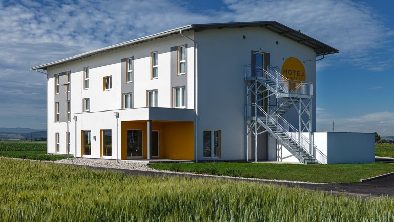 Exterior view of Hotel Tullnerfeld, © Hotel Tullnerfeld Exterior view of a modern hotel with a white façade and yellow accents, surrounded by fields.