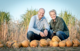 Two people kneel smiling behind a row of pumpkins in a field.