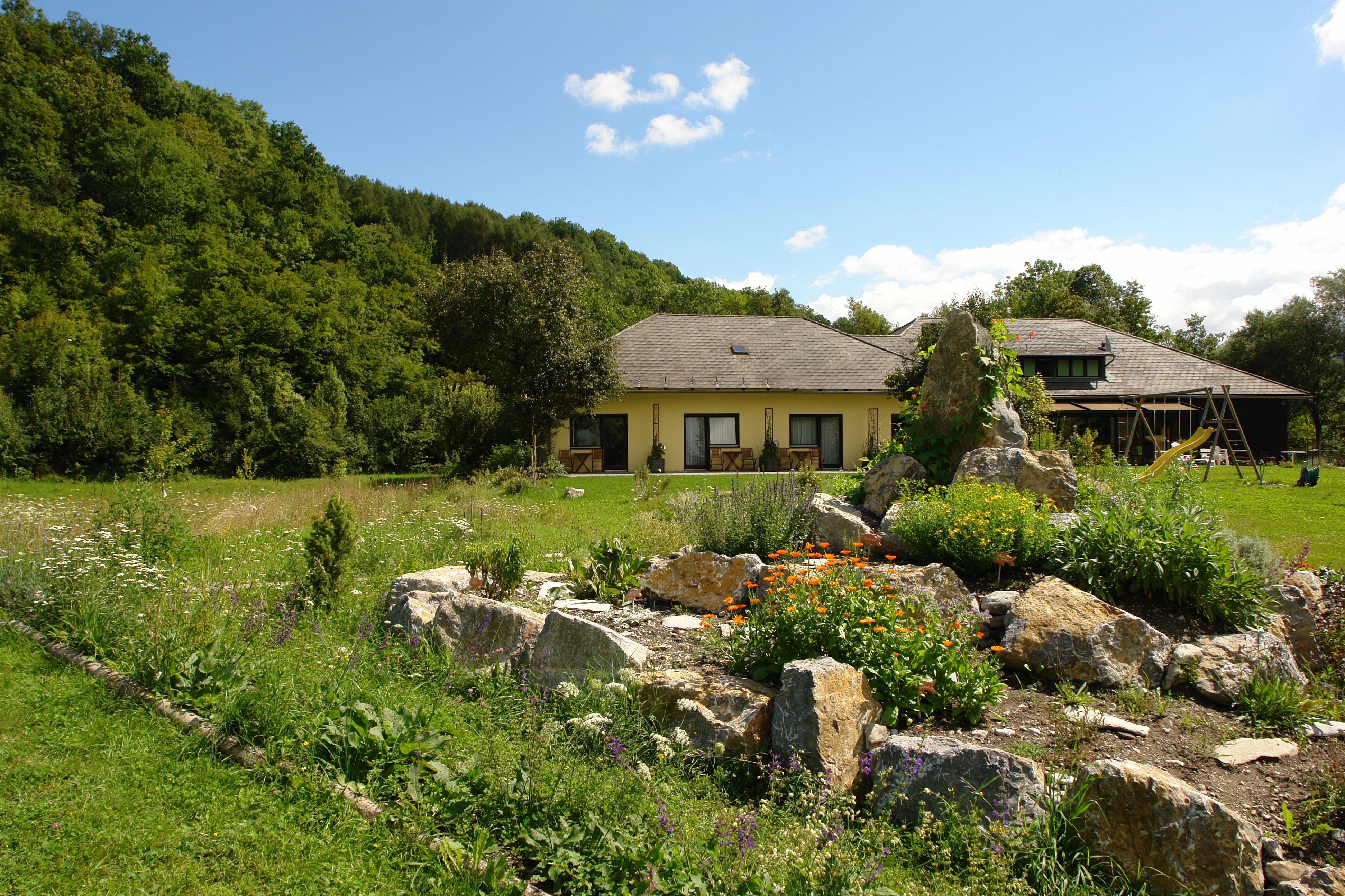 A yellow house with a garden and rocks in the foreground, surrounded by trees.