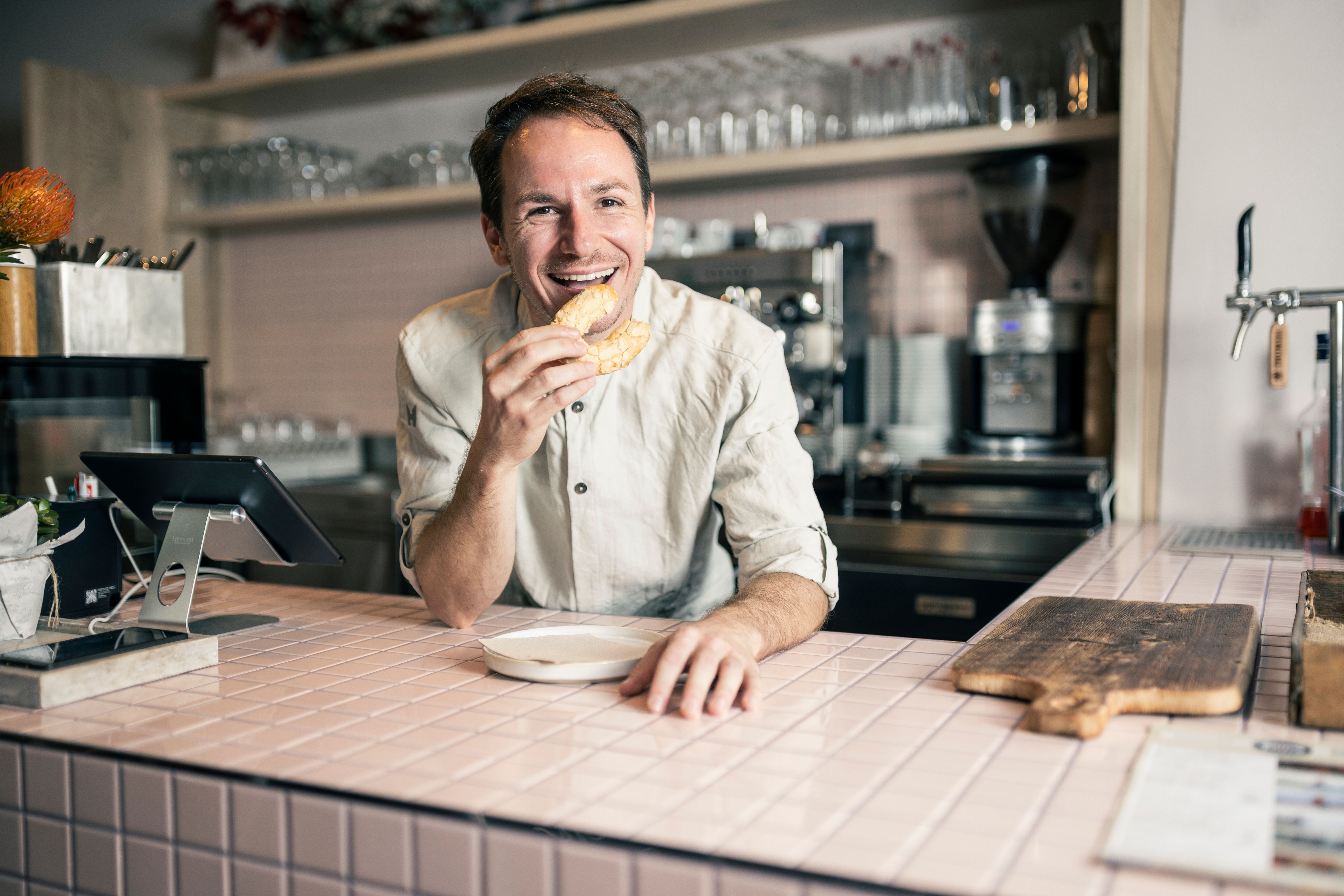 In einer einladenden Feinbäckerei genießt ein fröhlicher Mann einen frisch gebackenen Genuss. Die warmen Farben der Einrichtung und die köstlichen Backwaren schaffen eine gemütliche Atmosphäre, die zum Verweilen einlädt.