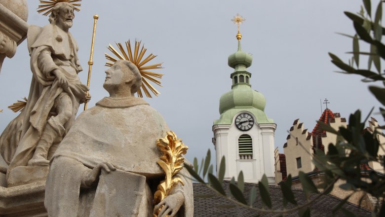Statue with halo in front of church tower with clock and green roof.