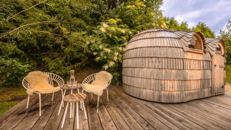 Wooden sauna with two chairs and table on a wooden terrace surrounded by greenery.