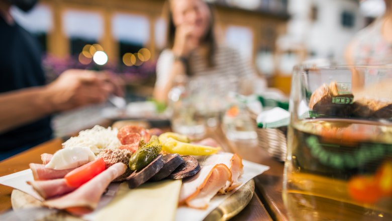 Close-up of a snack with sausage, cheese and vegetables on a table, blurred people in the background.