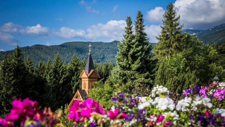Church tower surrounded by trees and flowers in front of a mountain landscape.