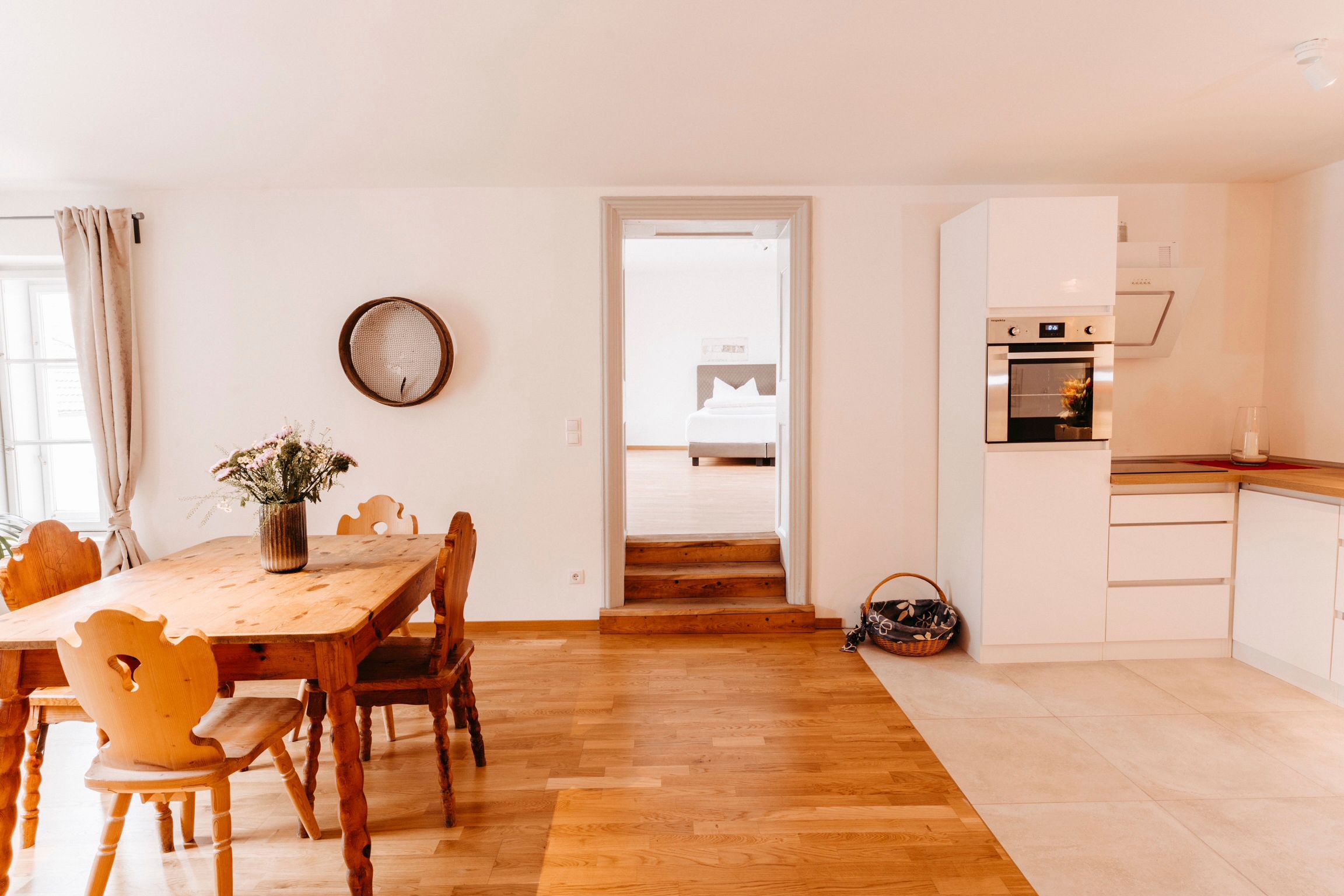 Bright kitchen with wooden table, chairs and modern white cupboards. Passageway to a bedroom in the background.