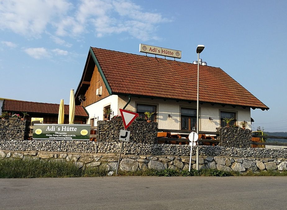 A small building with the inscription 'Adi's hut' and a red tiled roof, surrounded by stone walls and plants.