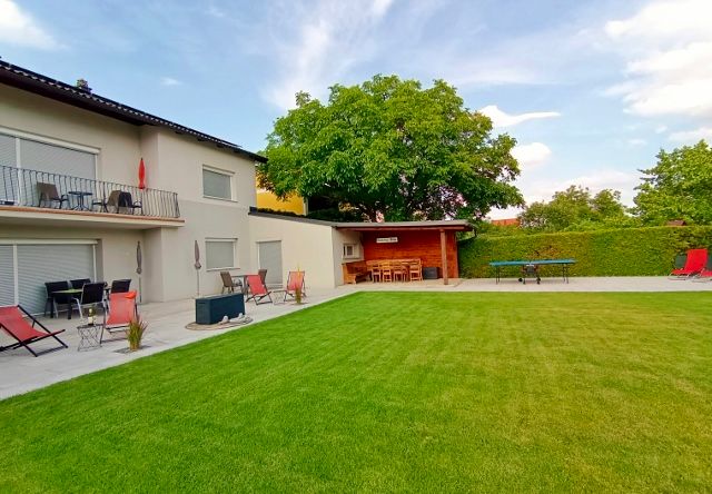 A well-kept garden with lawn, terrace, sun loungers and a large tree next to a modern building.