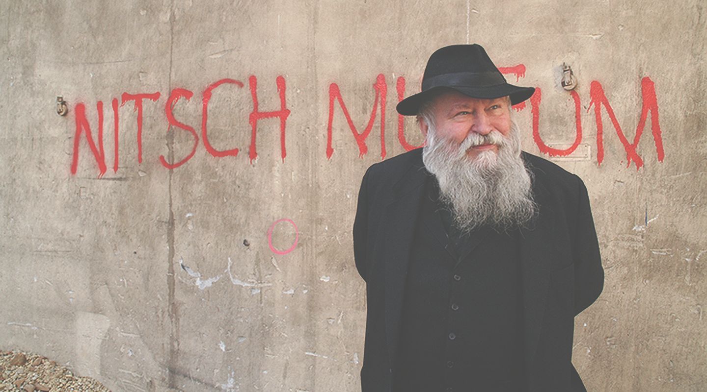 A man with a beard and hat stands in front of a wall with the inscription 'Nitsch Museum'.