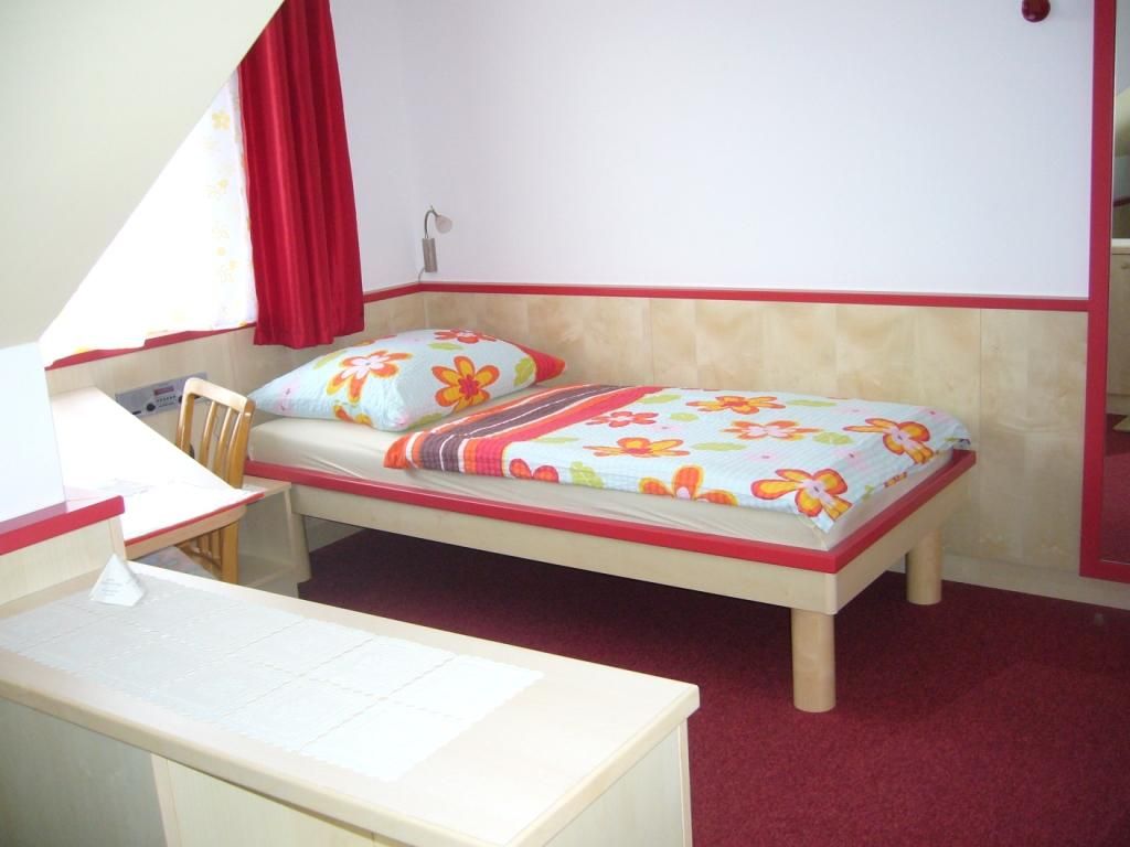 A simple room with a single bed, a desk and a chair. The bed linen has a floral pattern.