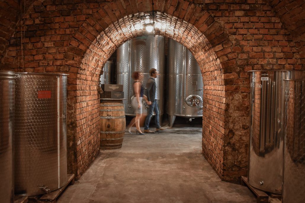 A wine cellar with brick walls and stainless steel tanks. Two people walk through a brick archway.