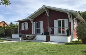 A red wooden house with a white roof and shutters, surrounded by a well-tended garden.