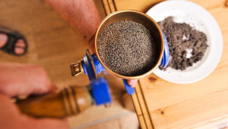 Close-up of a hand operating a mill with poppy seeds, next to it a plate with ground poppy seeds.