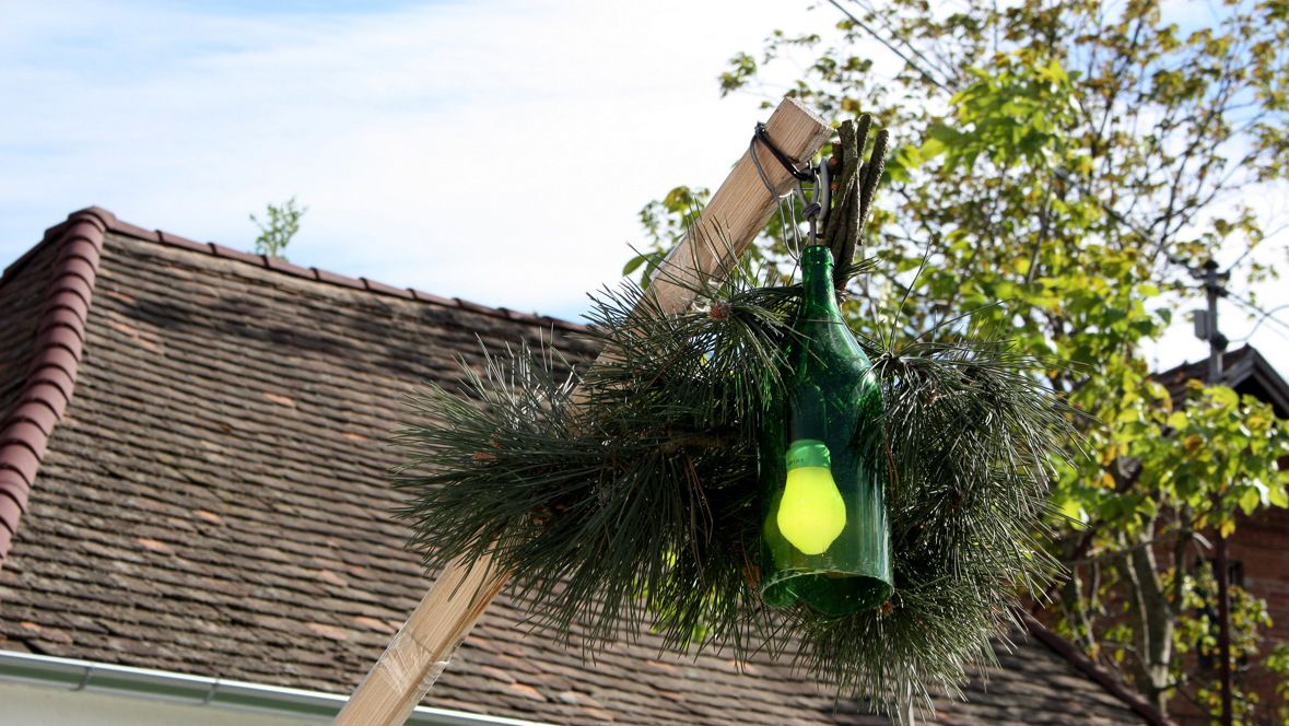 A green bottle with a yellow light bulb hangs on a wooden pole, surrounded by fir branches, in front of a roof and trees.