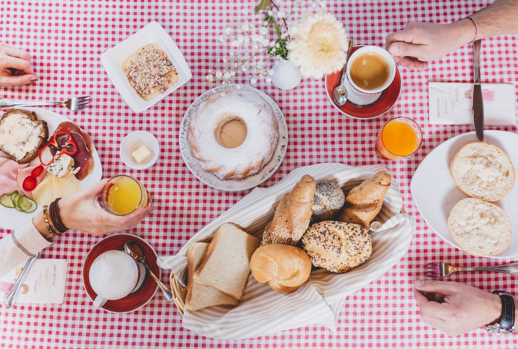 Breakfast table with bread rolls, cakes, cold cuts and drinks on a red and white checkered tablecloth.