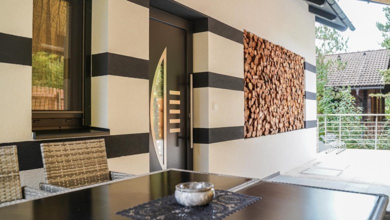 Entrance to a vacation home with a modern door, stacked wood on the wall and a table with chairs in the foreground.