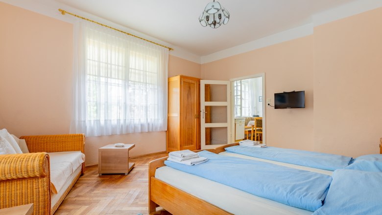 A cozy bedroom with two single beds, a sofa, a wardrobe and a small table. A window with curtains lets in natural light.