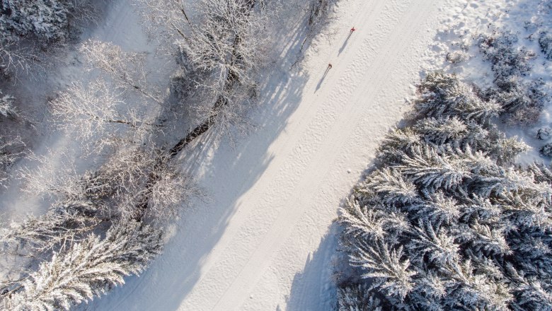 Aerial view of a snow-covered cross-country ski trail next to a stream with two people cross-country skiing, surrounded by snow-covered trees.