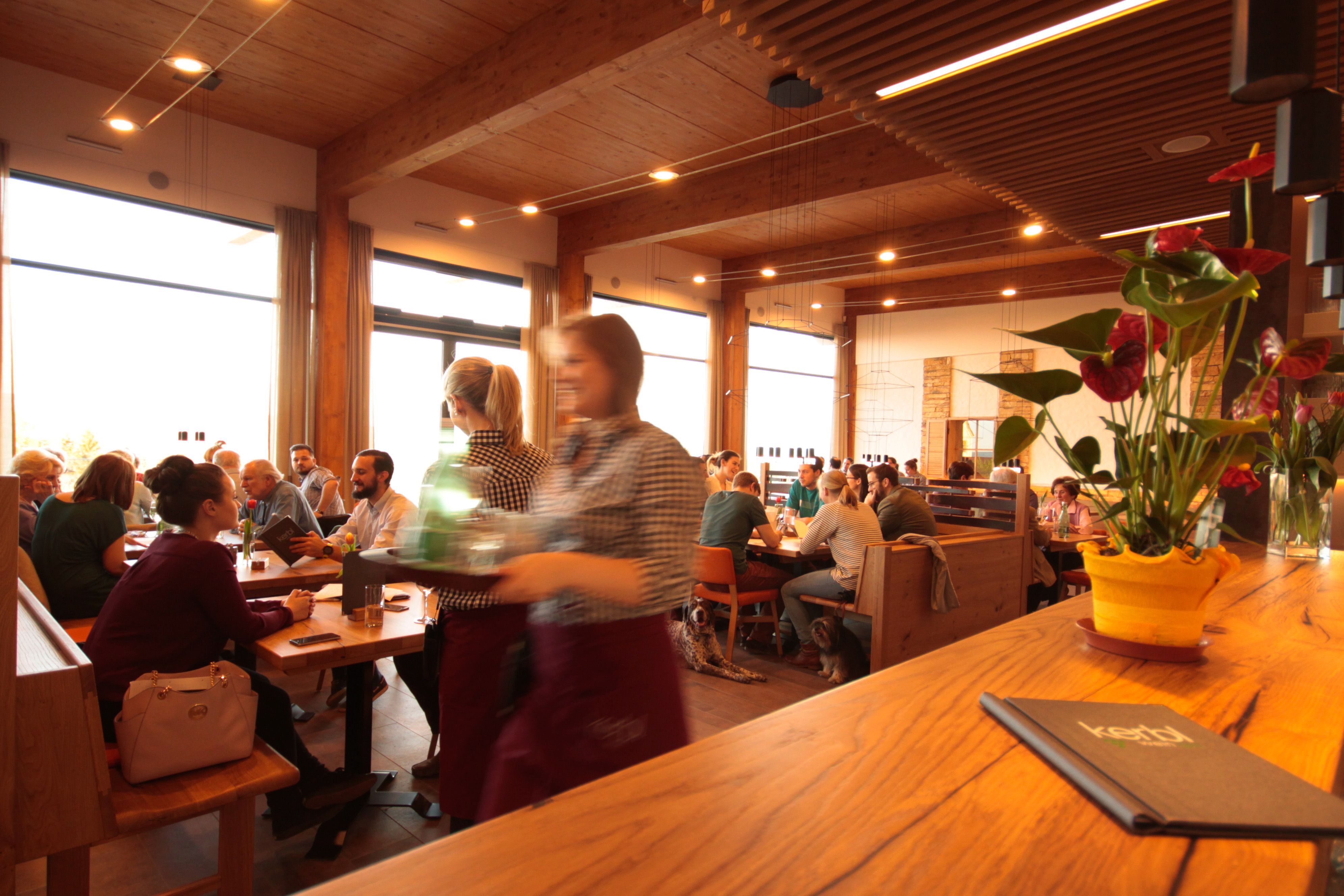 A busy dining room with wooden furniture, guests at tables and a waitress in the foreground. A plant stands on the bar.
