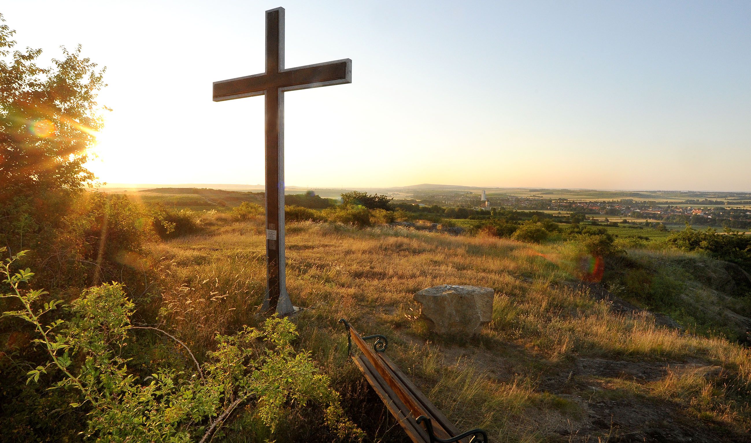 A cross on a hill at sunset with a view of a wide landscape and a village in the background.